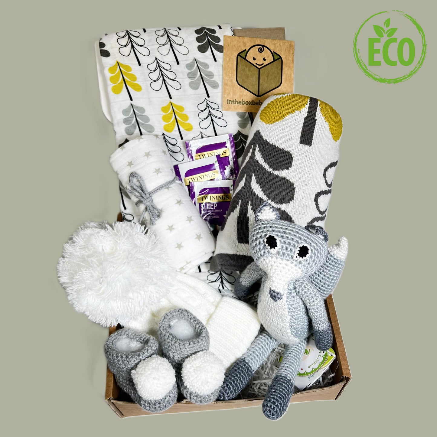 A neutral new parent gift hamper containg a lovely coton baby blanket and matching baby mulin, a smalle grey and white muslin, a white pompom babybhat, a pair of crocheted baby bootees in grey with white pompoms and a Sindibaba knitted fox rattle.