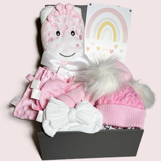 A pink and white new baby girl hamper in a grey magnetic baby keepsake box containing a pink and white cotton hooded baby towel with a giraffe motif fcae, a pink and white baby pompom hat, a pink cottob cellular baby blanket, a white baby headband, a pink Taggie blanket and a nursery sign