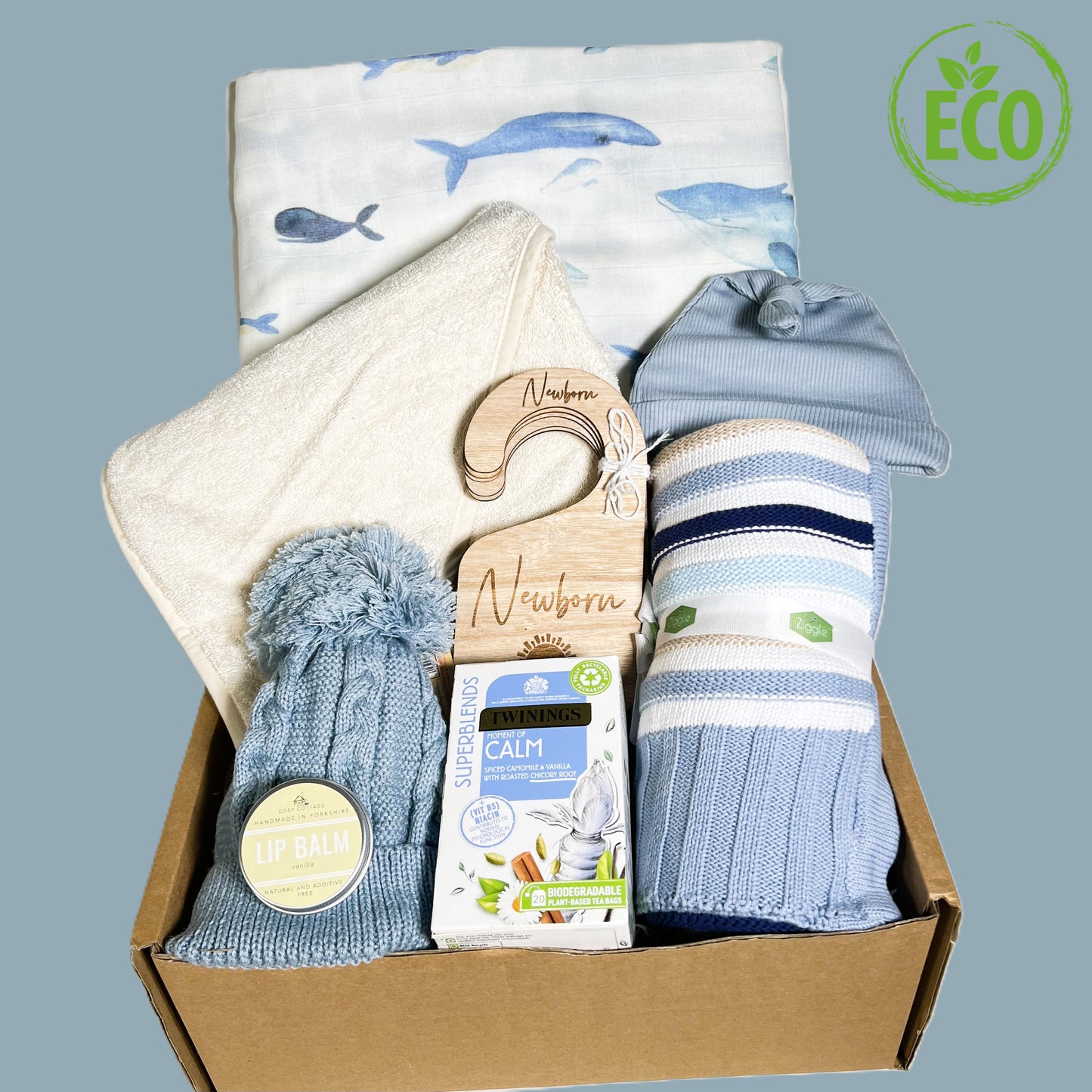 A blue new baby boy gift hamper cowitha blue white and beige cotton baby blanket, a large hale print baby msulin/swaddle blanket, an organic cotton hooded bby towel, a blue pompom baby hat, a blue cotton knotty hat, a set of woodedn baby wardrobe dividers, a vegan vanilla lip balm and a packet of Twining "Calm "teabags.