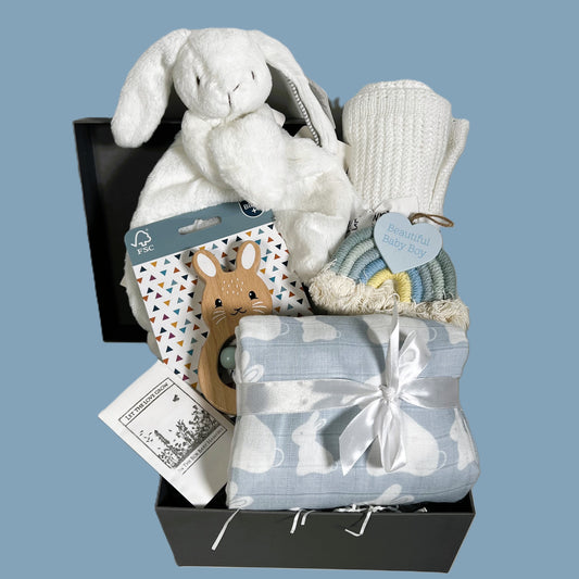 A baby boy hamper gift in  baby keepsake box containing A blue and white bunny print muslin swaddle blanket, a Mary Meyer bunny baby comforter, a white cellular baby blanket, a FSC wooden baby rattle and a nursery decoration.