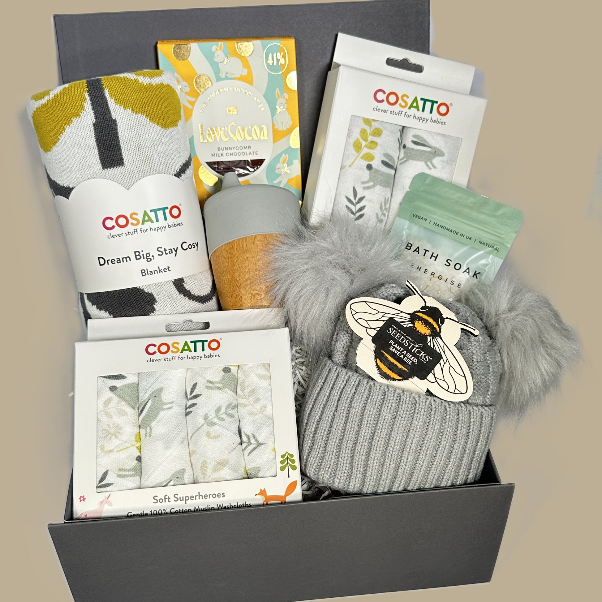 Baby gift hamper with Cosatto products including blankets, socks, and bath soaks on a beige background.