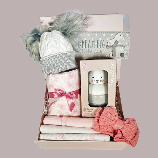 New baby gifts hamper with a pink rose bud baby blanket with cotton waffle to reverse and three matching baby muslins. A Rosa and Bo bunny stacking toy, a pink baby headband, a nursery plaque and a grey and white baby pompom hat.