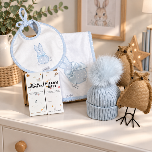 A curated baby boy hamper arranged on a white dresser, featuring a light blue and white bunny bib, a matching baby blanket, two boxed products labelled Bath & Massage Oil and Pillow Mist, a light blue knit beanie with a fluffy pom-pom.