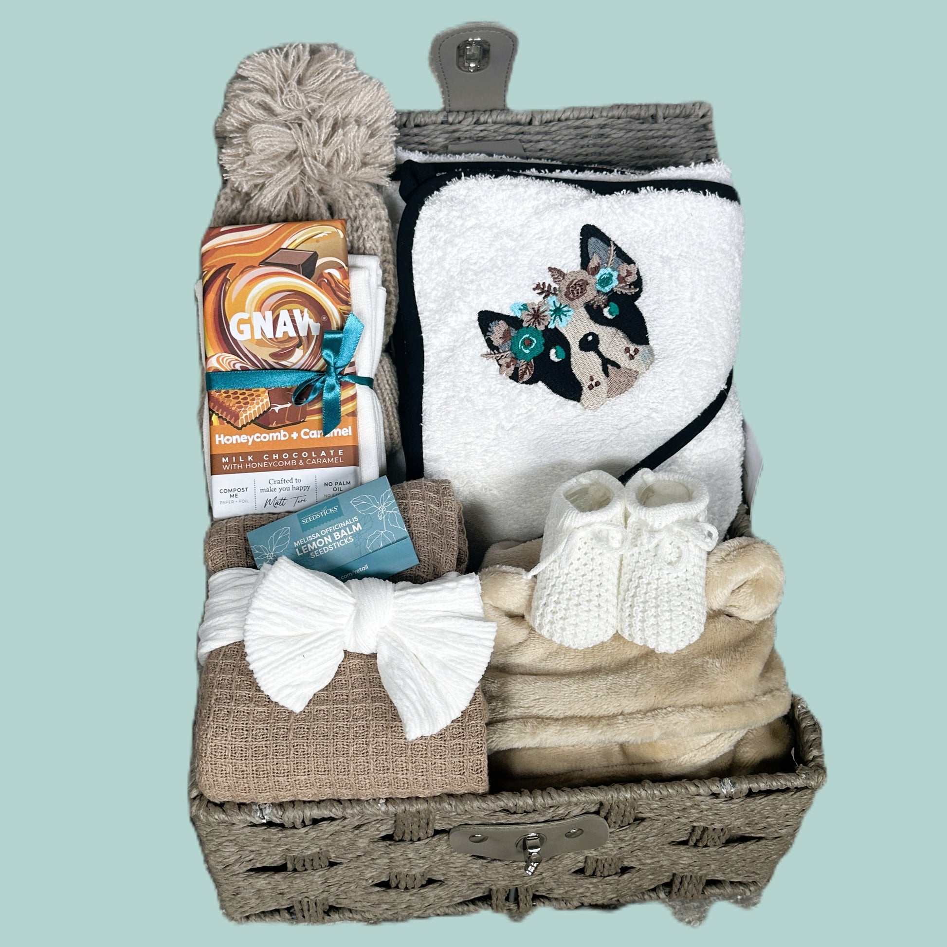 New Baby girl Gift basket with a towel featuring a French bulldog design, a box of chocolate and other items on a light blue background.