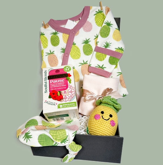 New baby girl hamper containing a organic cotton baby romper with pink, yellow and green pineapple print, matching baby knot hat, a crocheted baby rattle shaped like a pineapple and a box of Twinings teabags.