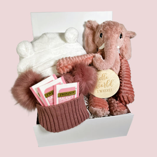 Pink baby girl hamper with a Pink cuddly elephant soft baby toy, a white hooded baby dressing gown, a dusky pink double pompom baby hat, Twinings teabags, a "Hello World" baby photograph sign and a white baby keepsake box.