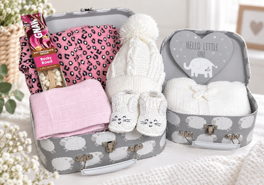 A baby gift set arranged in  grey baby keepsake cases with sheep print, alongside a matching heart-shaped nursery plaque. The set includes a white knit pom-pom hat, white bunny-face baby booties, a pink leopard-print sleepsuit, a folded pink muslin, Rocky Road hot chocolate spoon.