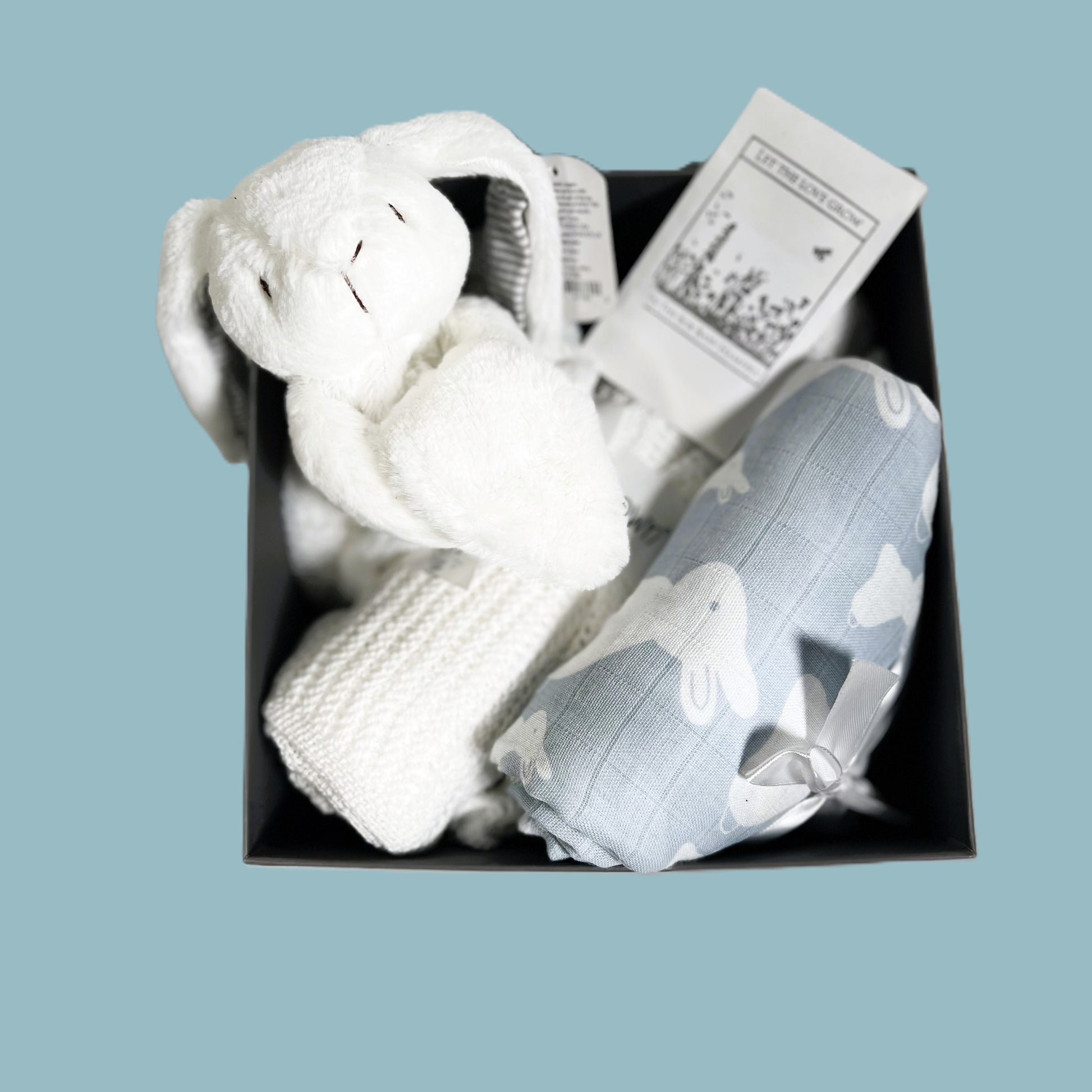 A baby boy hamper gift in  baby keepsake box containing A blue and white bunny print muslin swaddle blanket, a Mary Meyer bunny baby comforter, a white cellular baby blanket, a FSC wooden baby rattle and a nursery decoration.