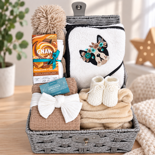 A baby gift basket arranged in a grey woven hamper, containing a white plush towel with a French bulldog dog embroidery, white baby booties, folded coffee coloured waffle baby blankey, a white baby headband, , a GNAW honeycomb chocolate bar, and a beige pom-pom baby hat and a taupe baby robe, set on a wooden surface with soft home decor in the background.