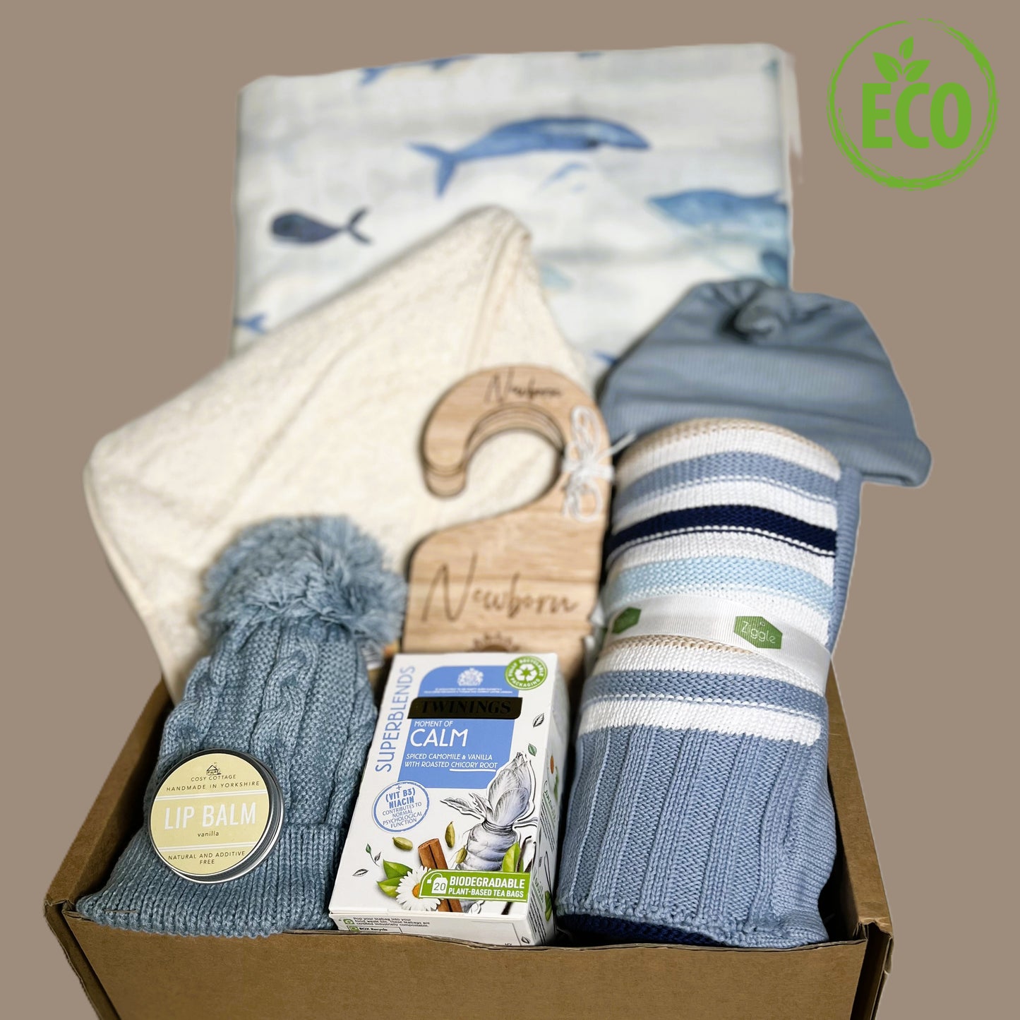 A blue new baby boy gift hamper cowitha blue white and beige cotton baby blanket, a large hale print baby msulin/swaddle blanket, an organic cotton hooded bby towel, a blue pompom baby hat, a blue cotton knotty hat, a set of woodedn baby wardrobe dividers, a vegan vanilla lip balm and a packet of Twining "Calm "teabags.