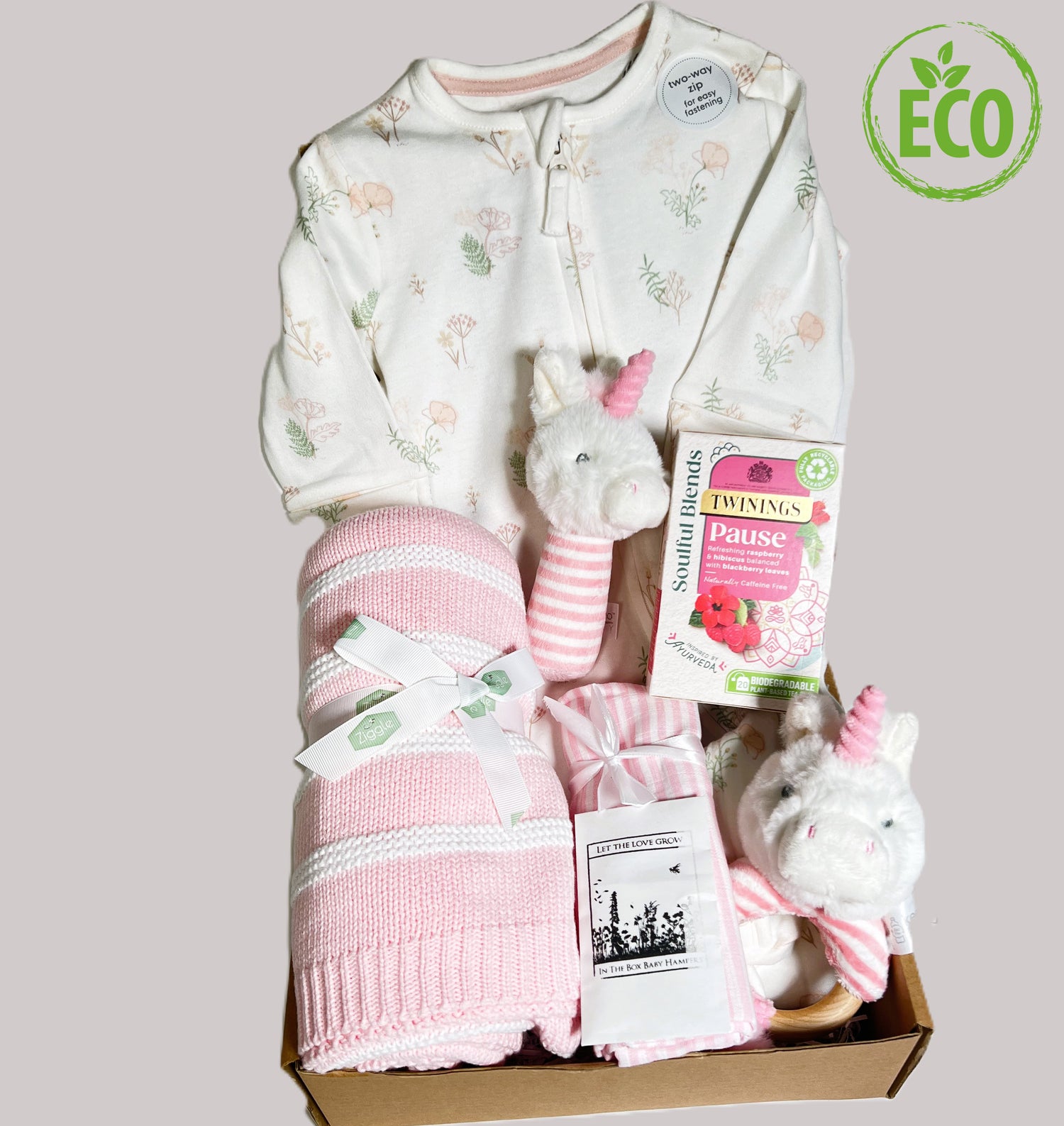 New baby girl gift hamper containing an organic cotton zipped baby sleepsuit white with a pink and green flower print, a white and pink striped cotton baby blanket, 2 Keeleco unicorn baby rattles, a muslin square and a pack of Twinings Pause teabags.