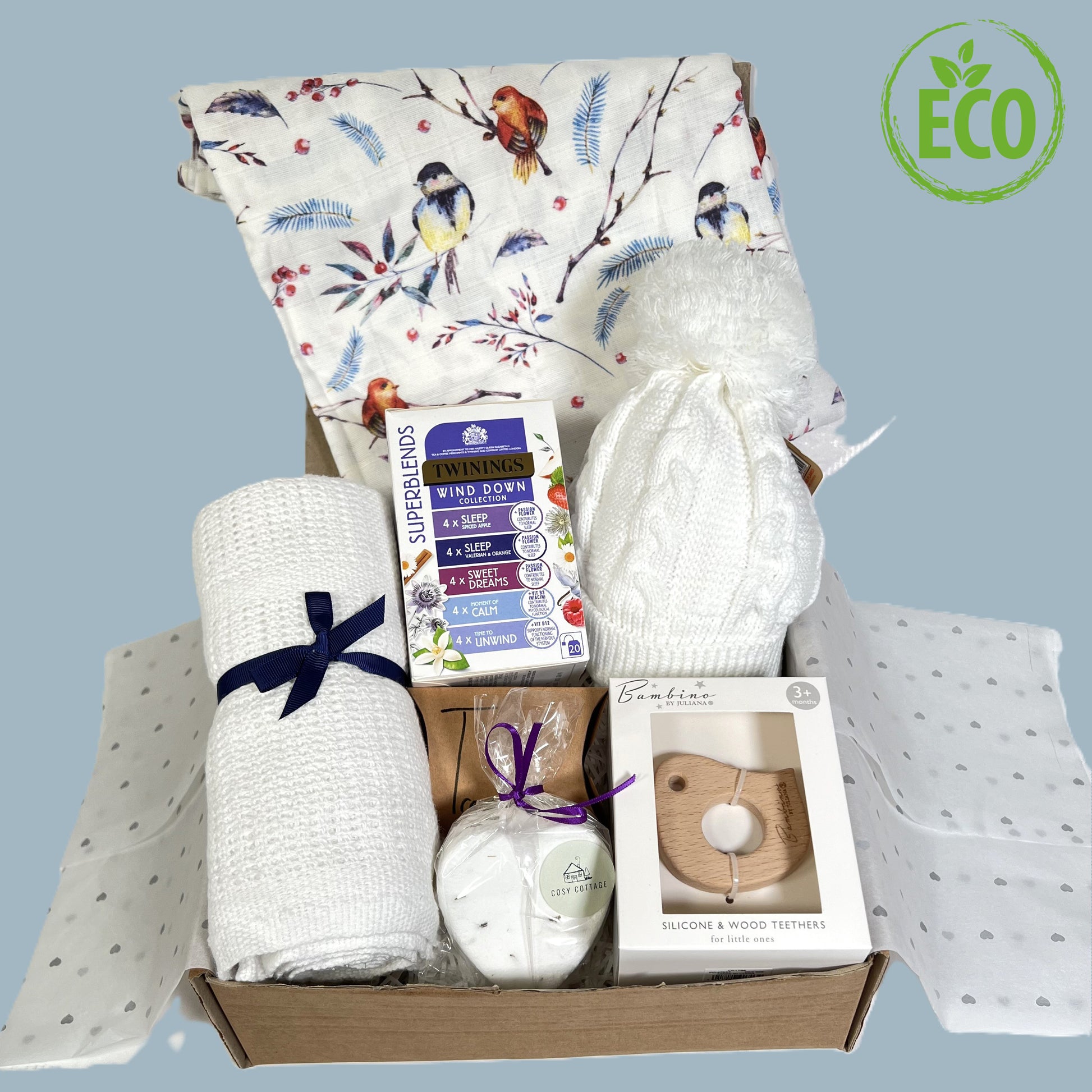 Eco friendly newborn baby girl gift includes a large whie baby muslin with a bird print, a white cottoncellular baby blanket, a baby pompom hat, a wooden bird baby teether, a heart shaped bath bomb and some sleepy tea for the new parents.