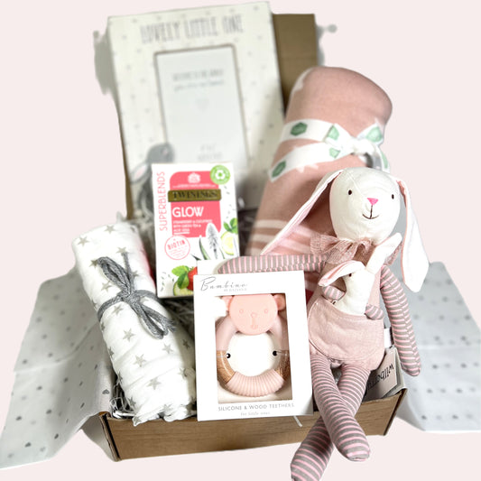 A new parents gift hamper for a baby girl includes a pink star print cotton baby blanket, a bambino bear teether, a white and grey muslin square, a wooden photo frame and a packet of "Glow" teabags.