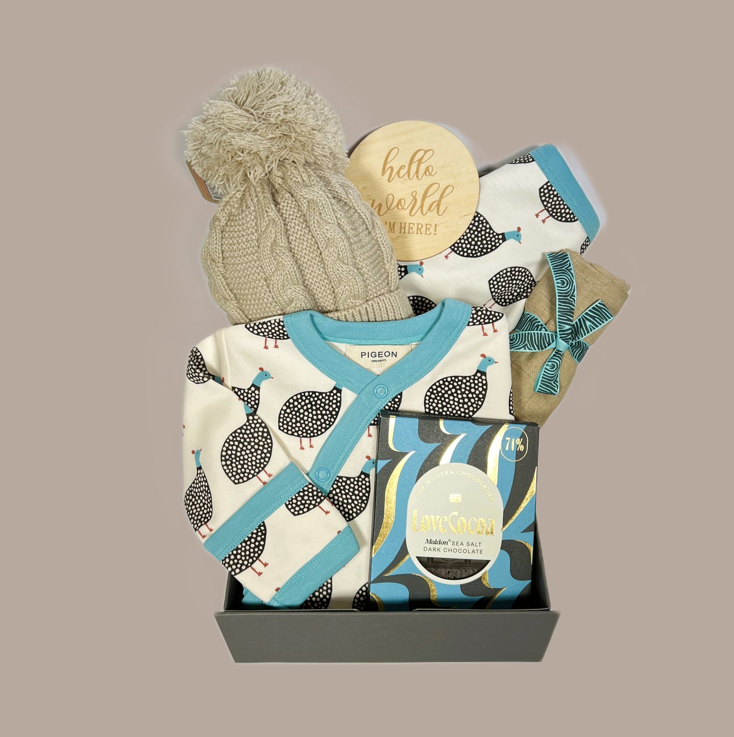 A baby shower gift in a magnetic gift box containing an organic baby sleeps suit in a Guinea fowl print with matching reversible baby dribble bib, A bar of Love Cocoa Malden sea salt dark chocolate, a taupe baby muslin and a taupe baby pompom hat.