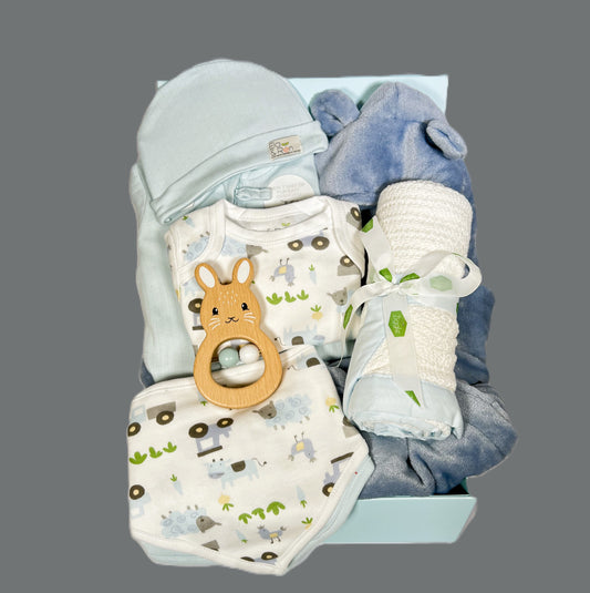 A new baby boy hamper containing Organic cotton baby layette set with double zipped baby sleepsuit, 2 baby dribble bibs, a baby hat and a farmyard print short sleeved baby body suit. A which cotton cellular baby blanket with a blue cotton trim, a wooden bunny baby rattle and a dusky blue baby dressing gown.