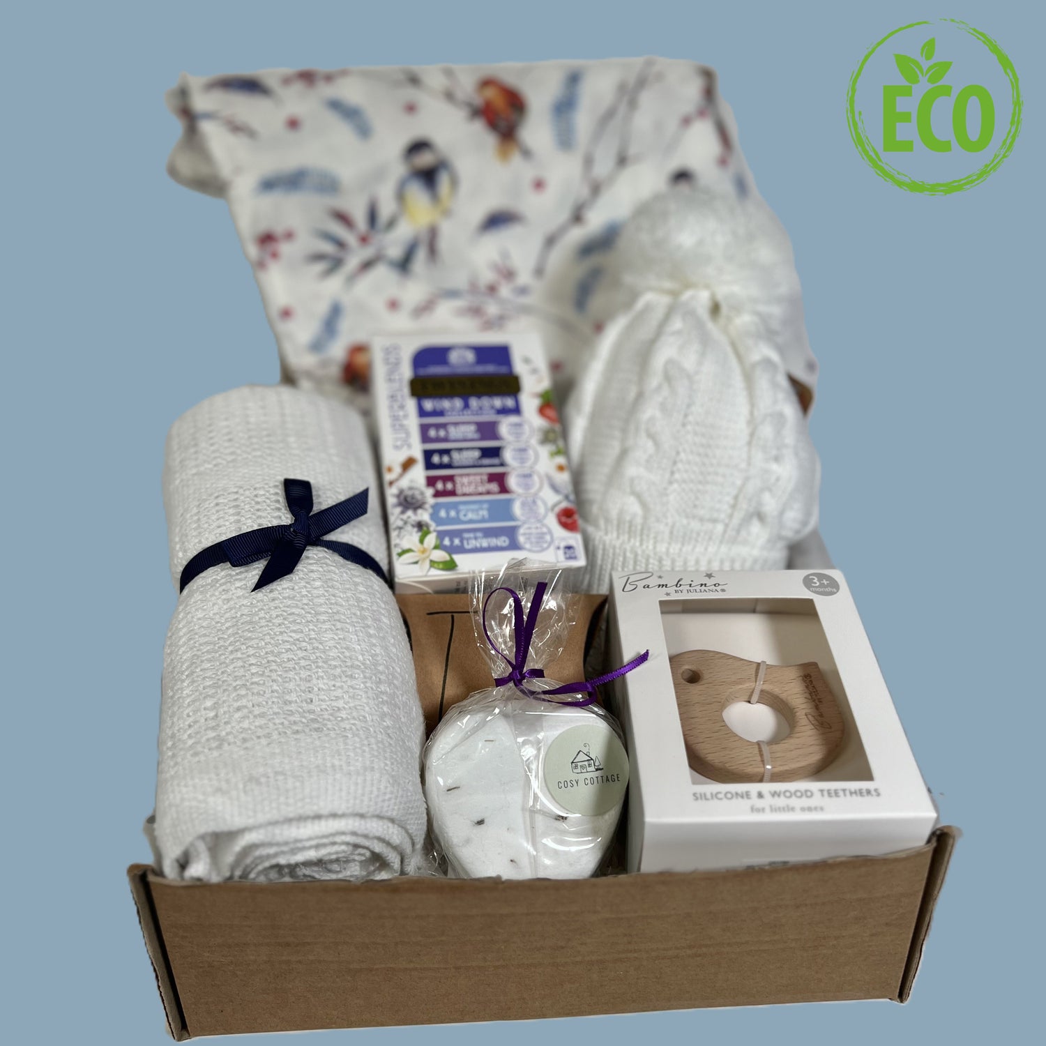 Eco friendly newborn baby girl gift includes a large whie baby muslin with a bird print, a white cottoncellular baby blanket, a baby pompom hat, a wooden bird baby teether, a heart shaped bath bomb and some sleepy tea for the new parents.