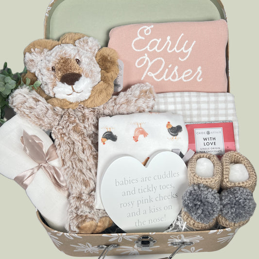 New baby girl gift containing organic cotton baby bodysuits in peach , one reading " Early Riser". A Mary Meyer Lion lovey, a pair of hand crocheted baby booties with pompoms, a mslin square, a "with love " bar of chocolate and a nursery plaque, all in a kraft baby keepsake case with an abstract white flower print.