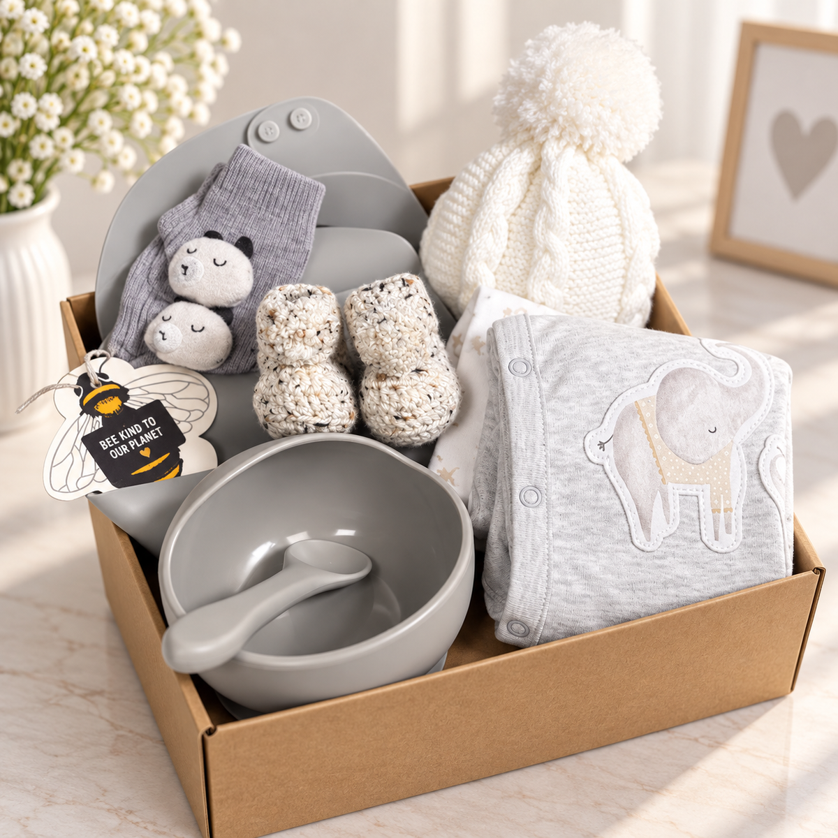 A curated neutral new baby hamper with silicone baby bib, feeding bowl and spoon, 2 long sleeved baby body suis, a pair of chrocheted baby booties with pompom, a white baby hat and some baby socks.