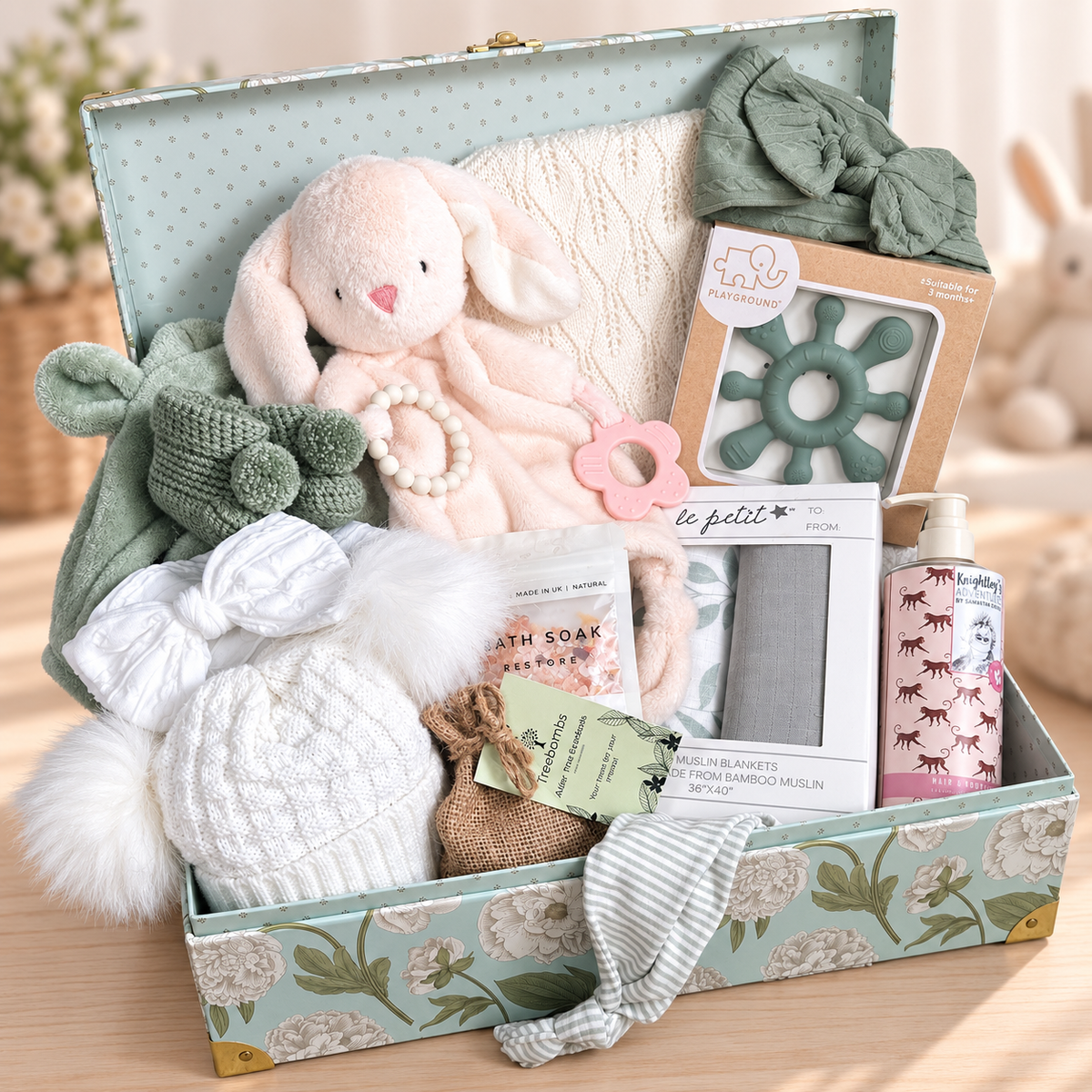 A baby gift box filled with soft baby items, including a plush bunny, knitted baby blanket, a white baby pompom hat, baby wash, bath salts, crocheted baby booties, baby robe in a sage colour pink baby comforter with silicone teething rings, arranged in a floral mint green box on a wooden surface.