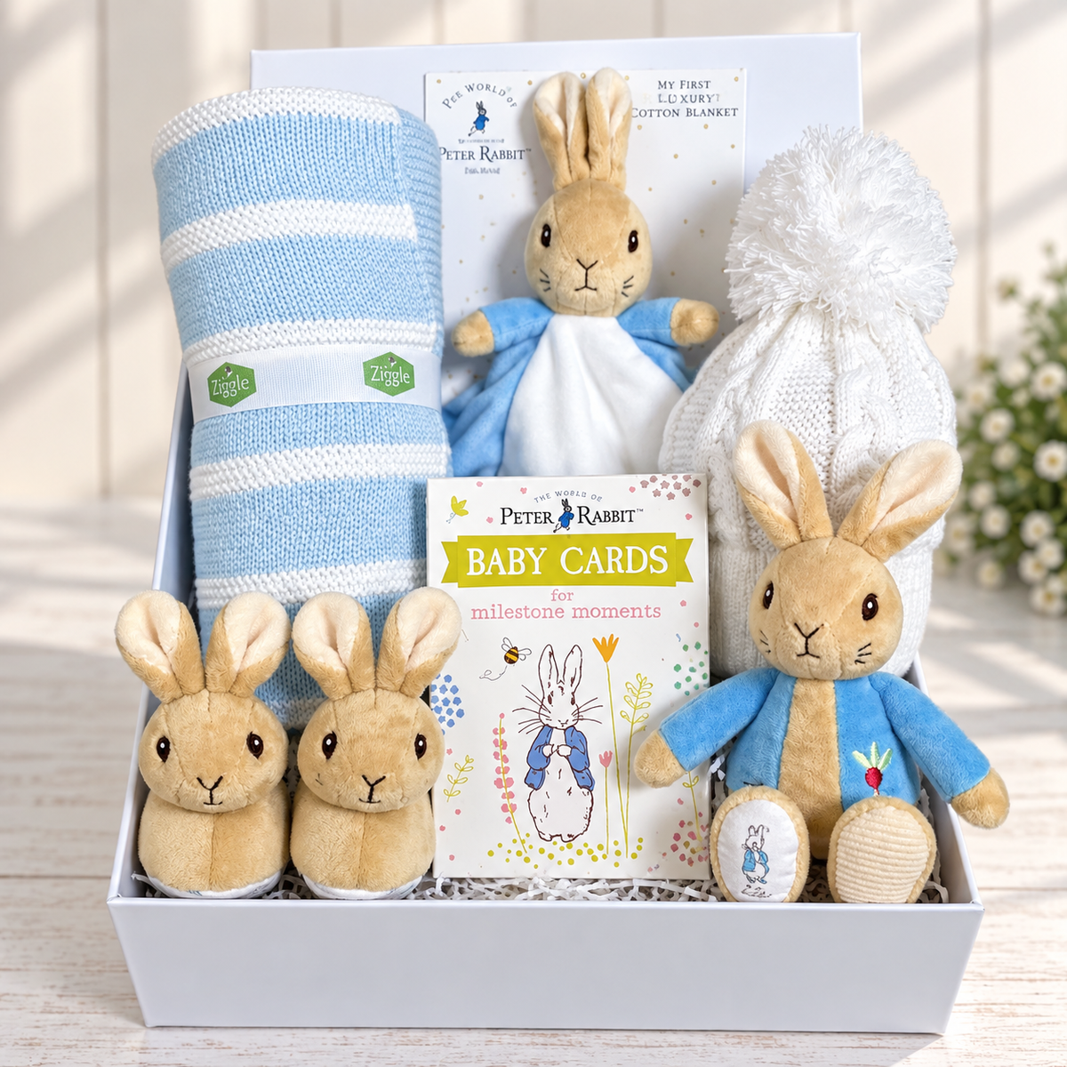A curated Peter Rabbit baby gift set arranged in a white baby keepsake box, including a blue-and-white striped blanket, Peter rabbit baby comforter and baby rattle, a white knit baby  hat with pom-pom, and Peter Rabbit baby milestone cards. 