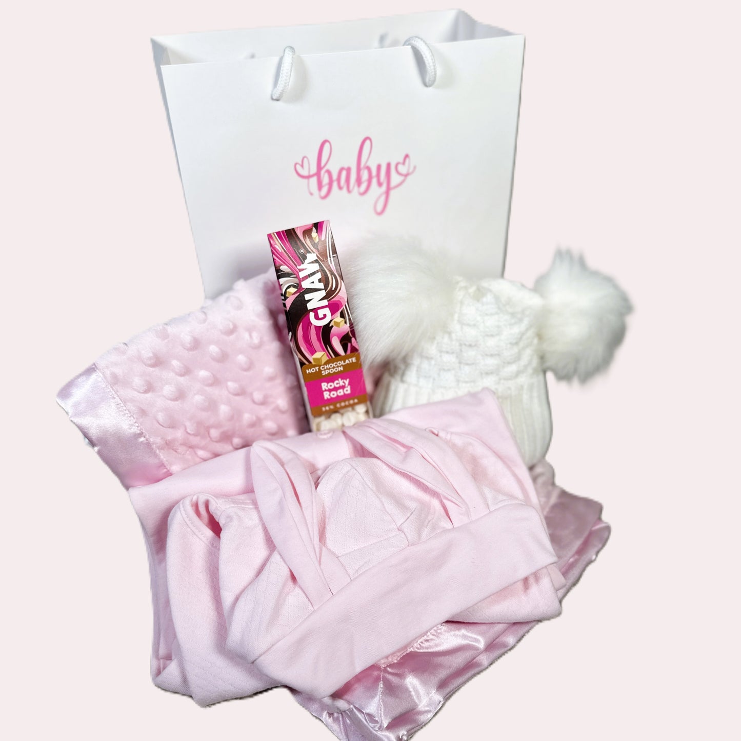 Baby gift with pink blanket, hat, and product on a Gnaw rocky road hot chocolate stirrer white background.