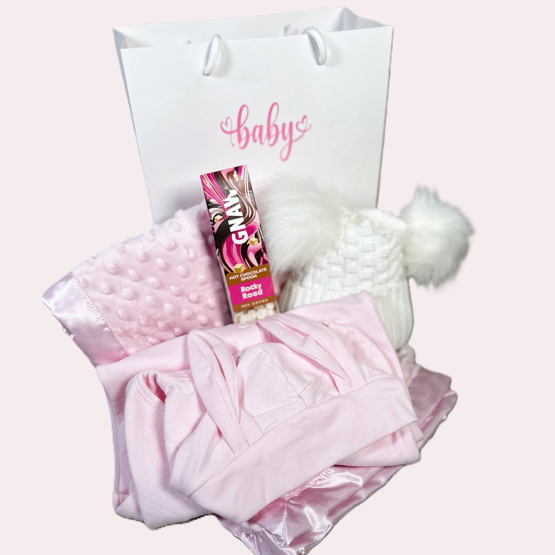 Baby gift with pink blanket, hat, and product on a Gnaw rocky road hot chocolate stirrer white background.