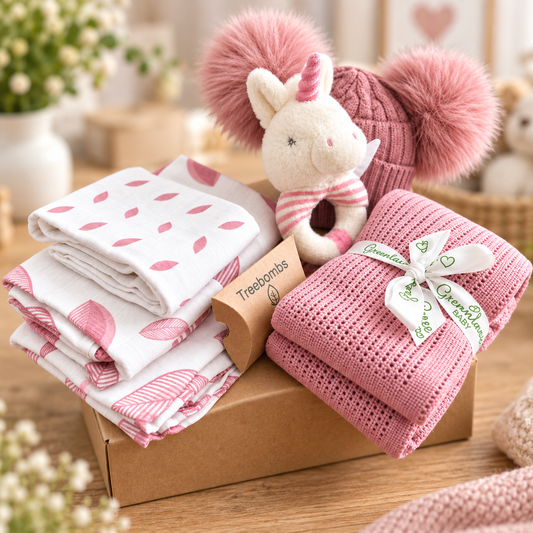 A curated rose pink and white baby girl hamper gift, featuring a pink knit baby pompom hat with, 3 baby muslins in rose pink leaf print, a pink cellular baby blanket tied with a white ribbon, and a Keeleco unicorn baby rattle.