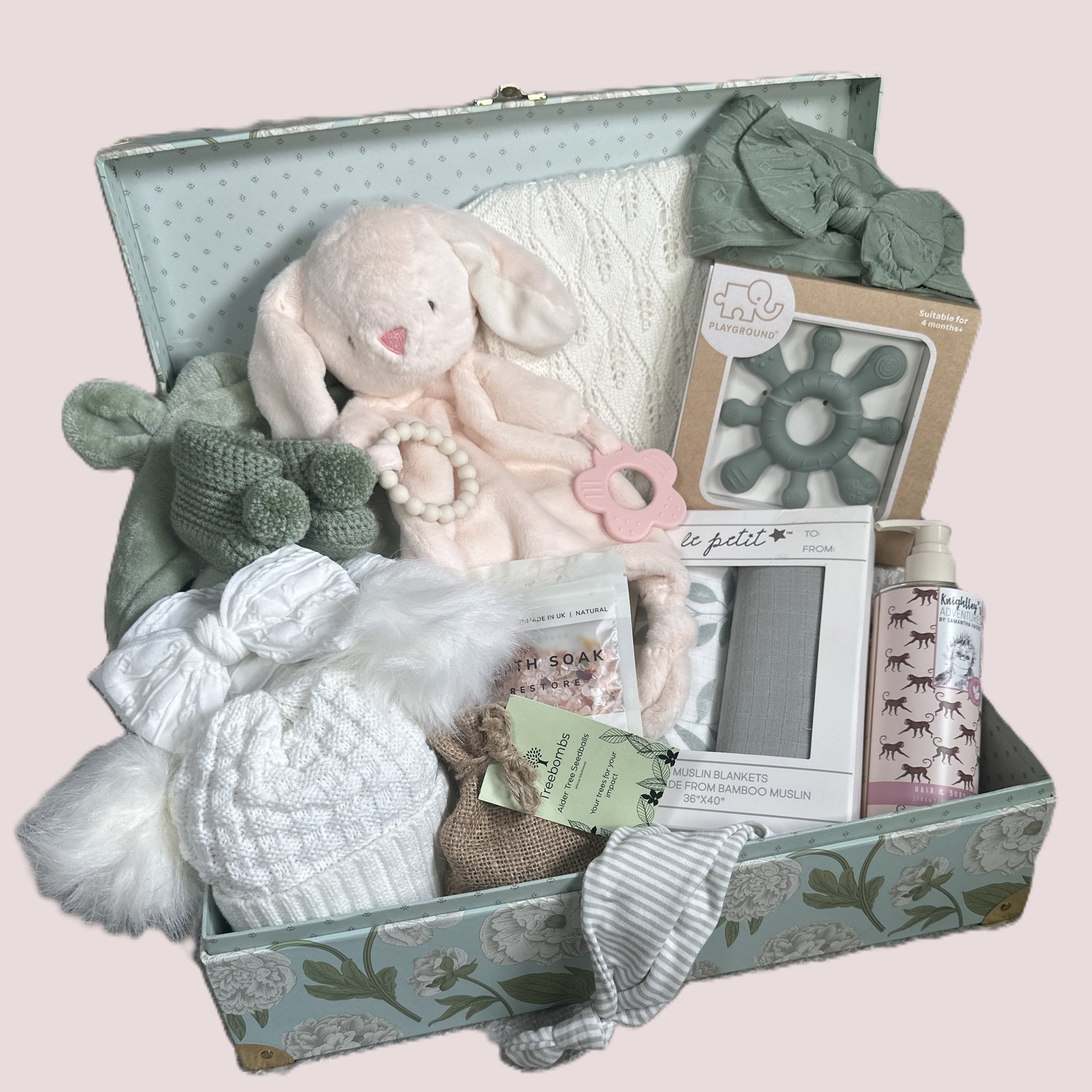 Gift box with baby items including a plush toy, knitted items, and baby care products on a light pink background.