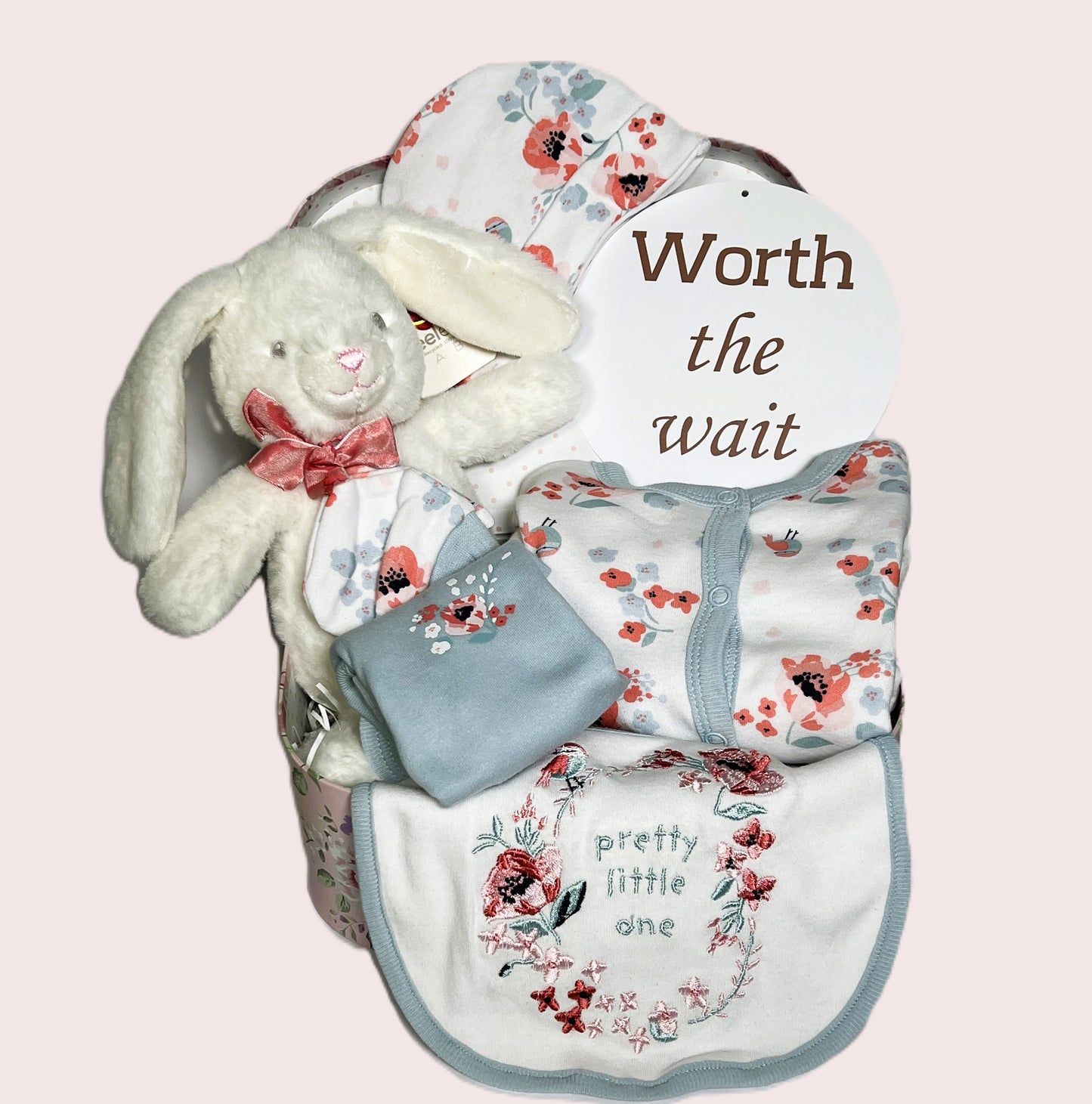 A baby girl hamper in a floral baby keepsake case containing a Keeleco soft bunny baby toy and a white pink and blue baby layette set including a baby sleep suit, a baby body suit, a matching hat and a pair of scratch mitts.