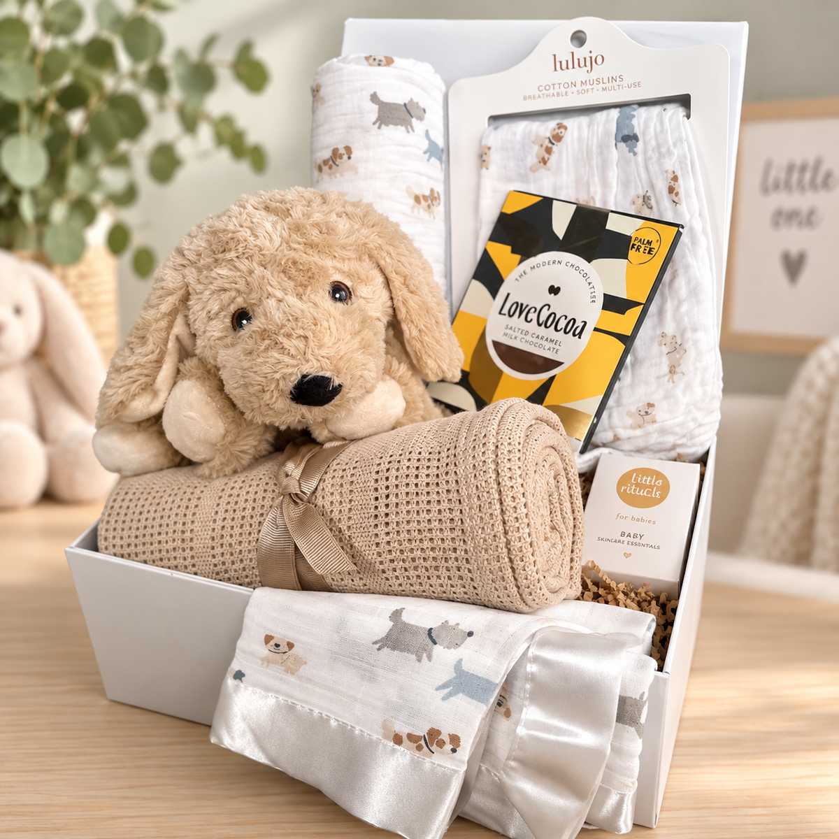 A  unisex new baby gift hamper in a white box, featuring a plush tan puppy toy, a Taupe cellular baby  blanket, white muslin swaddle  blanket with dog prints, , Lulujo cotton swaddle blankets with the same puppy print and a satin edged comforter , styled on a wooden table with a blurred nursery background.