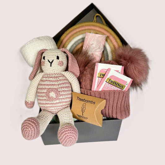 A new baby girl hamper in a grey baby keepsake box containing  a crocheted bunny baby soft toy, a white cotton baby blanket, a rose pink baby pompom hat, some Twinings teabags and a pink and white muslin square.