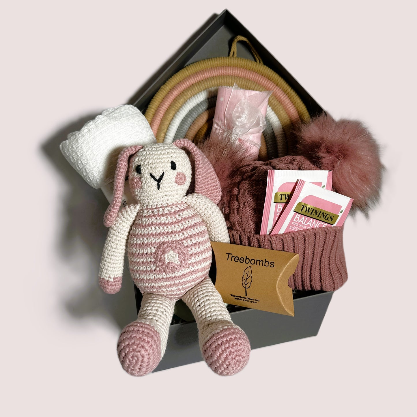 A new baby girl hamper in a grey baby keepsake box containing  a crocheted bunny baby soft toy, a white cotton baby blanket, a rose pink baby pompom hat, some Twinings teabags and a pink and white muslin square.