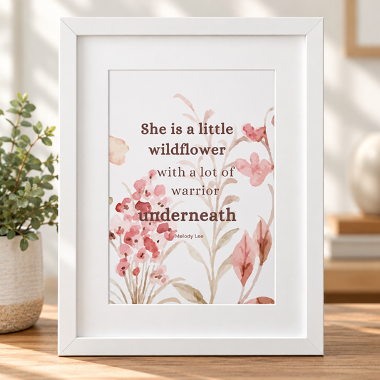 An A4 white-framed art print featuring a quote by Melody Lee, 'She is a little wildflower with a lot of warrior underneath,' surrounded by pink watercolor flowers, displayed on a wooden surface next to a potted plant.