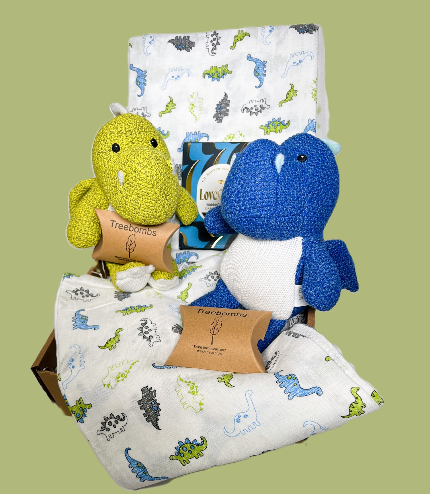 Twin boys baby gift hamper containing two Wilberry knitted dragon soft baby toys, 1 is blue the other is green, 2 large muslins with a dinosaur print in blue white and green, 2 packets of Treebombs and a bar of Love Cocoa chocolate.