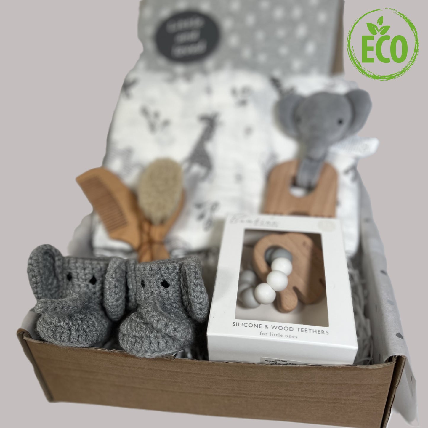 A stunning new baby hamper full of elephant baby essentials such aa a large white baby muslin with grey elephant and zebra print. Two elephant baby teething toys. A pair of crocheted baby booties with an elephant face, a grey organic cotton baby blanket and a baby hairbrush and comb set.