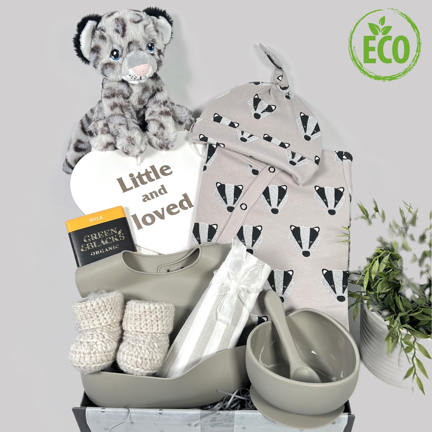 A luxury unisex new baby gift containing a GOTS certified organic cotton baby romper and matching baby knot hat in a stone colour with a badger face print, a weaning set in silicone, a pair of crocheted baby booties, aaaaaaakeeleco snowleopard soft baby toy, a bar of Green and Blacks chocolate and a \Nursery sign reading "Little and loved".