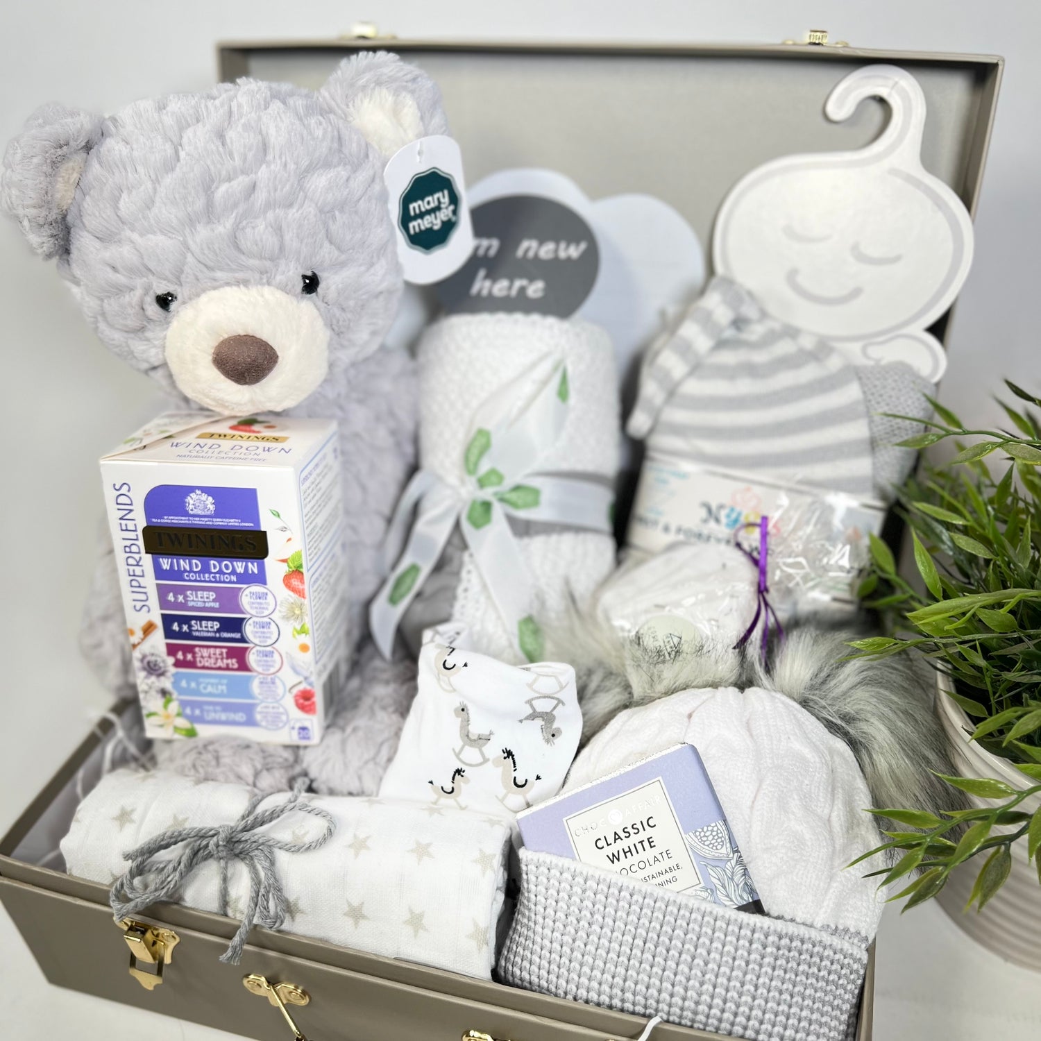  A neutral new baby gift in a khaki baby keepsake case containing 1 medium Mary Meyer shadow teddybear soft toy, a white cotton cellular baby blanket with a grey trim, 1 grey swaddle blanket and matching baby knot hat,a large musling, a cotton dribble bib, a grey and white baby pompom hat, a natural lavender bather bomb, a packet of Twining calming and sleep teabags and a bar of Choc Affair white chocolate.