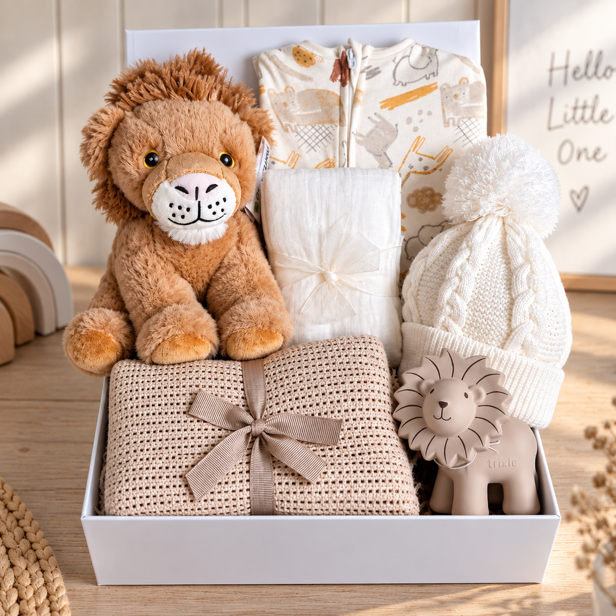 A curated baby hamper in a white baby keepsake box featuring a plush lion toy, a whit baby hat with a pom-pom, a taupe cotton cellular baby blanket tied with a ribbon, and a patterned baby outfit, A Tikiri baby teether and a white baby muslin arranged on a wooden surface.