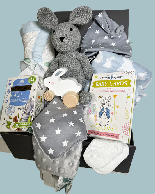 Blue and grey new baby boy hamper gift contining a white cellular cotton baby blanket with a blue trim,  a blue and white bunny print bamboo baby swaddle blanket, a gret Taggie blanket, a grey crocheted bunny soft baby toy with an embroidered face, a pack of Peter Rabbit milestone cards, a matching grey and white star print baby bib and baby knot hat and a pack of Twinings calming teabags.