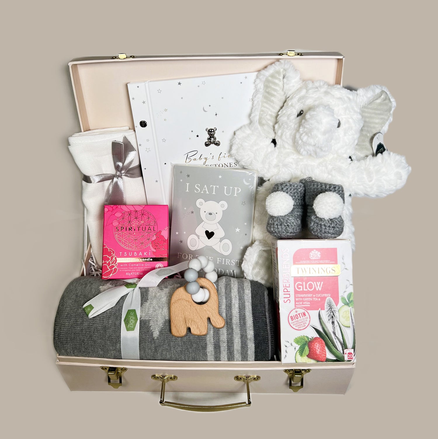 A Cream baby keepsake case containing a grey cotton baby blanket, a Mary Maeyer white elephant lovey soft baby toy, a soya scented candle, a pair of chrocheted baby bootees in dark grey with a white pompom, a wooden elephant shaped baby teether and a set of baby milestone cards and matching baby photo album