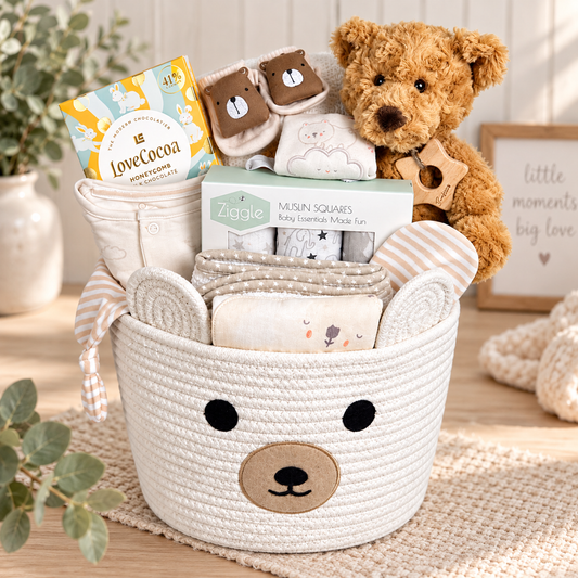 A baby gift basket set in a woven cream bear-face basket, filled with baby items including  Ziggle muslin squares, babywear, a hooded baby towel, baby socks, and a brown teddy bear holding a wooden toy.