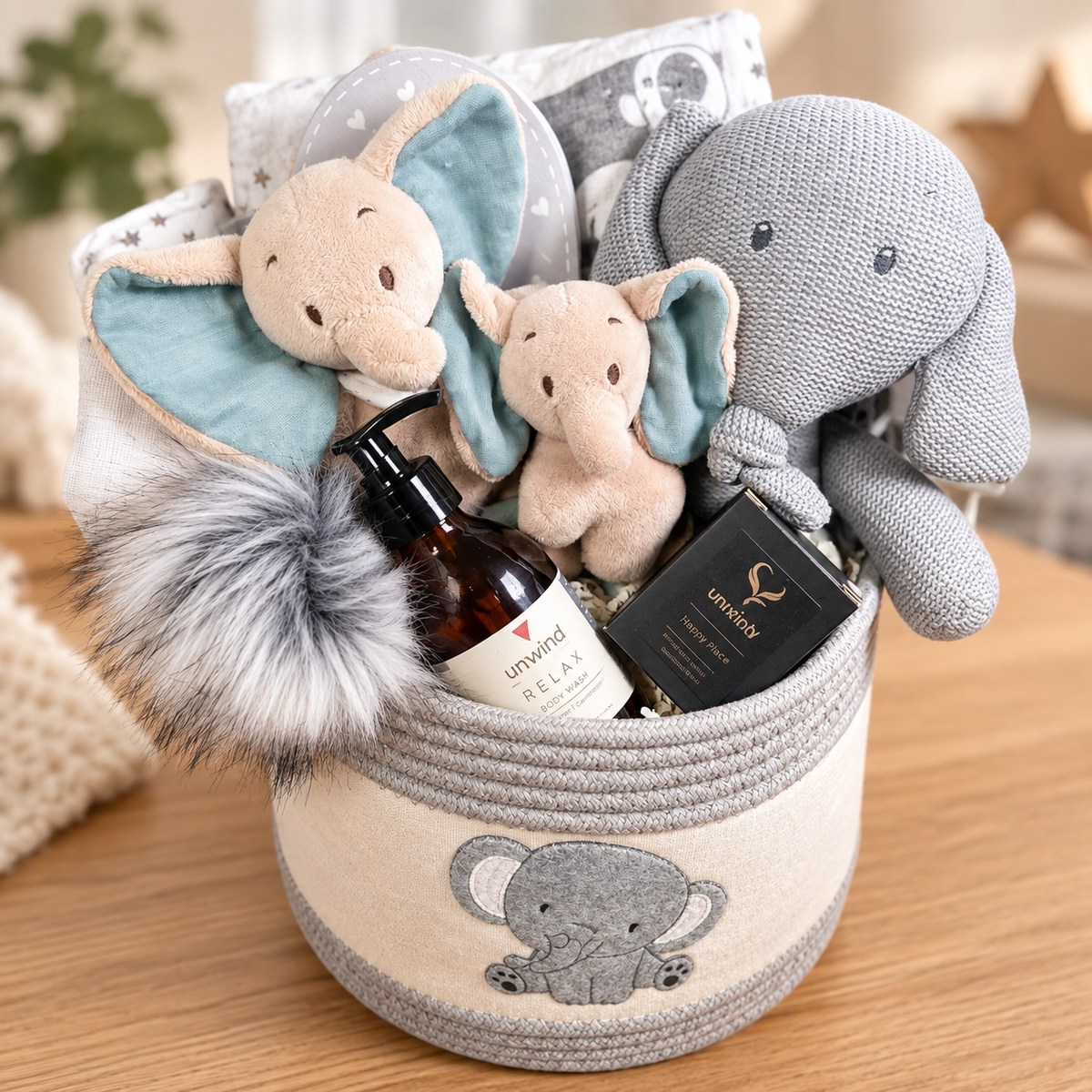 A curated new baby gift basket filled with plush elephant toys, a bottle of Relax shower gel, a black boxed item, and a grey Nattou Tembo elephant baby toy, arranged in a cream and grey rope basket on a wooden surface.