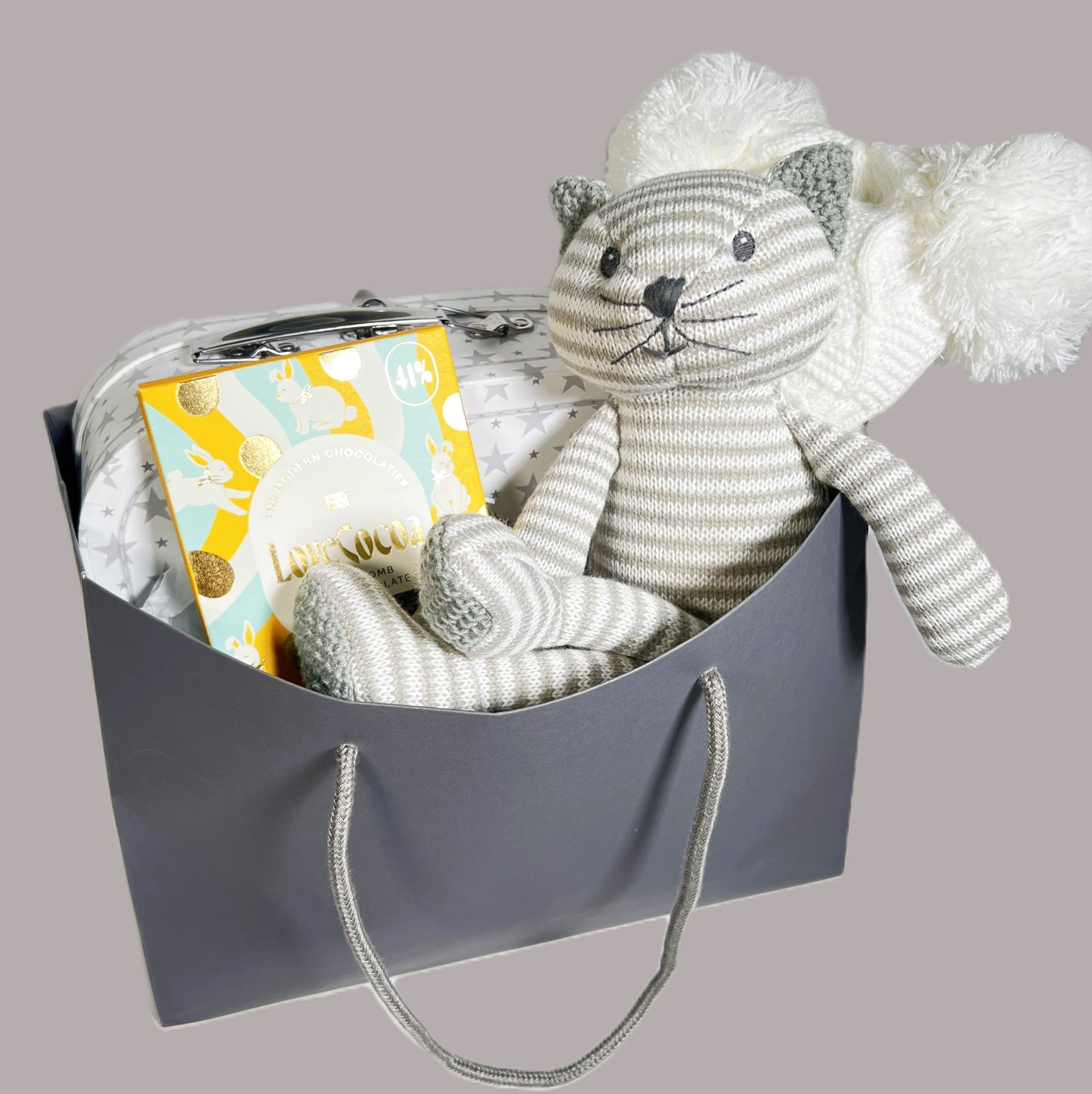 Neutral new baby gift in a bag containing a grey and white stripped knitted cat toy, a white baby pompom hat, a bar of Loce Cocao choclate, a kitty print baby layette set and a white storage suitcase with grey star print.