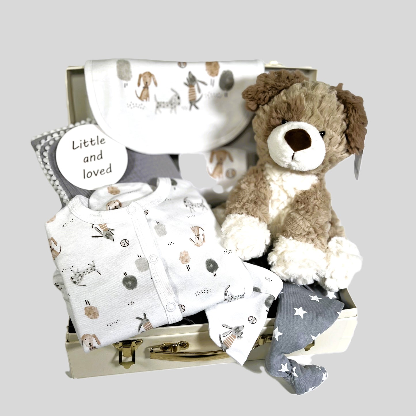 New parents baby gift in a cream baby keepsake case containing a cotton layette set withs a puppy print in neutral colours, a soft white baby blanket, a grey muslin square with pompoms and a Ziggle baby knot hat in grey with white stars.