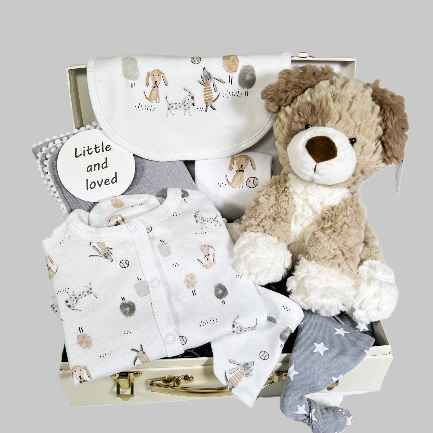 New parents baby gift in a cream baby keepsake case containing a cotton layette set withs a puppy print in neutral colours, a soft white baby blanket, a grey muslin square with pompoms and a Ziggle baby knot hat in grey with white stars.