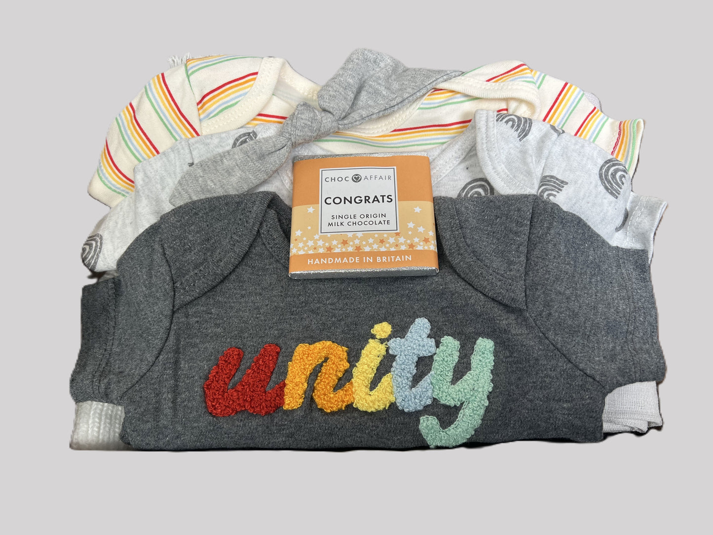 Unisex organic cotton new baby gift with 3 organic cooton baby bodysuits, a wooden elephant push along toy, a grey baby knot hat, a muslin square a baby pompom hat, a grey cellular baby blanket and a bar of chocolate.