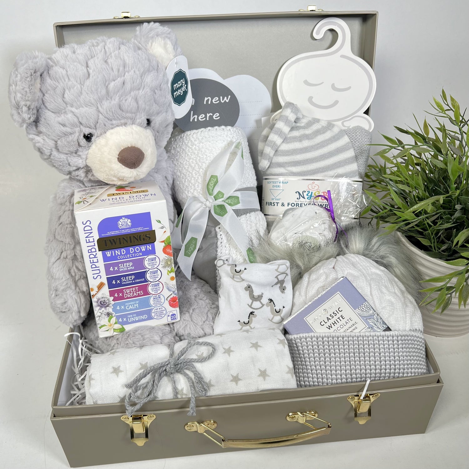 A neutral new baby gift in a khaki baby keepsake case containing 1 medium Mary Meyer shadow teddybear soft toy, a white cotton cellular baby blanket with a grey trim, 1 grey swaddle blanket and matching baby knot hat,a large musling, a cotton dribble bib, a grey and white baby pompom hat, a natural lavender bather bomb, a packet of Twining calming and sleep teabags and a bar of Choc Affair white chocolate.