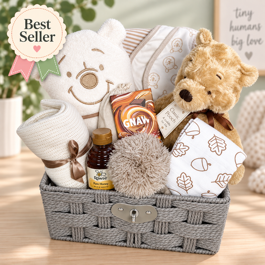 A baby gift basket arranged in a gray woven paper rope, filled with a plush winnie the pooh bear toy, Winnie the pooh hooded baby towel, patterned baby bibs, a rolled marshmallow colour cellular baby blanket with a brown ribbon, a small bottle of honey, a fluffy Taupe baby pom-pom, and a bag of GNAW chocolate. A 'Best Seller' badge appears in the top left corner.