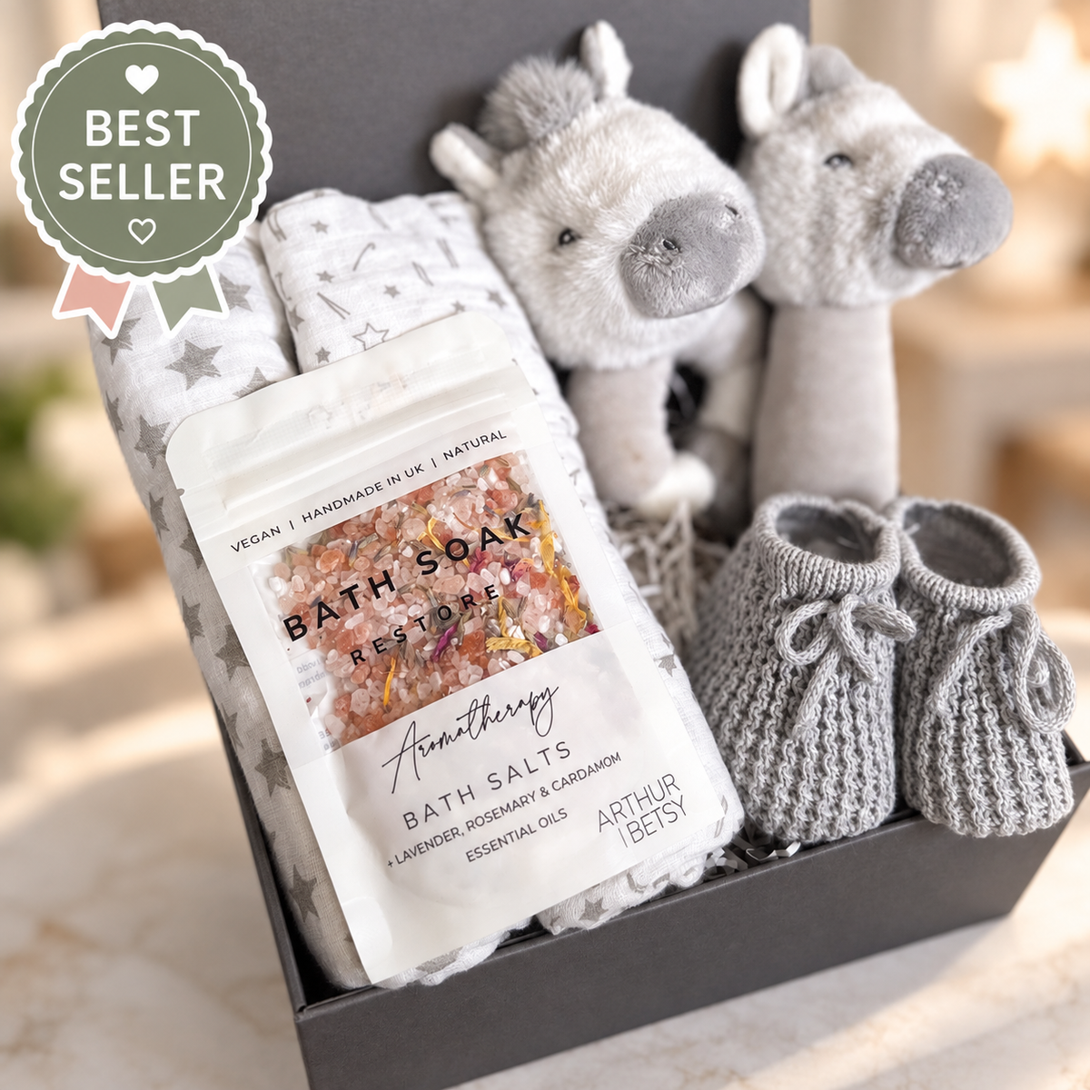 A baby gift set in a dark grey gift box featuring a bag of Arthur Betsy Bath Soak "Restore" bath salts, folded 70cms baby muslins, and a  Keeleco baby rattle, one round,  a stick rattle, a pair of grey knit booties. 