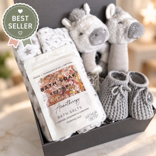 A baby gift set in a dark grey gift box featuring a bag of Arthur Betsy Bath Soak "Restore" bath salts, folded 70cms baby muslins, and a  Keeleco baby rattle, one round,  a stick rattle, a pair of grey knit booties. 